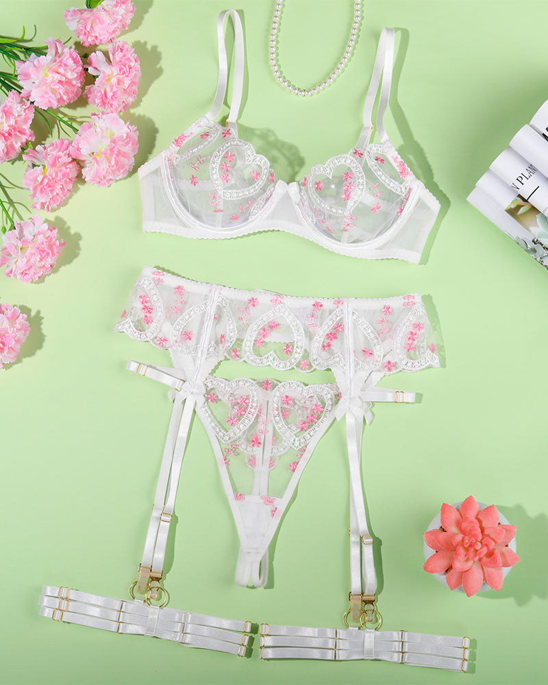 White heart-embroidered lace bra, matching garter thong, and thigh garters set, flat lay.
