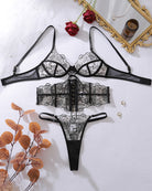 Black embroidered lace bra, corset belt and thong set laid out with accessories